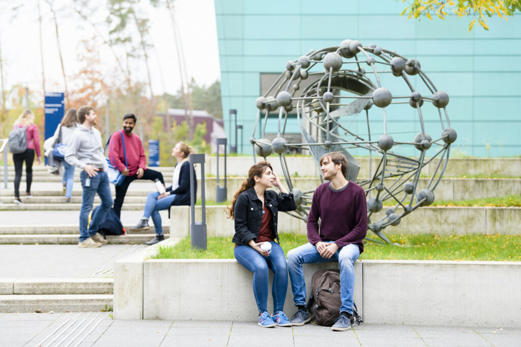 Two students are sitting in front of a piece of art outside.